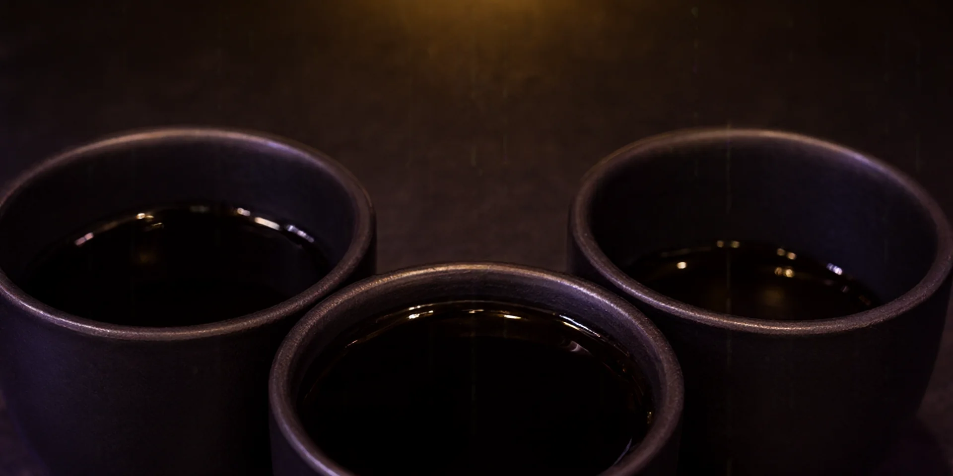 Tres pequeñas tazas de cerámica negra mate idénticas en formación triangular cada una con un nivel diferente de líquido oscuro sobre una superficie de piedra oscura.