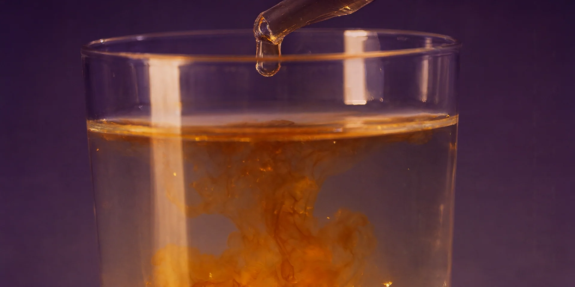 Une pipette libère une goutte ambrée dans un verre d'eau, et la couleur se diffuse lentement.