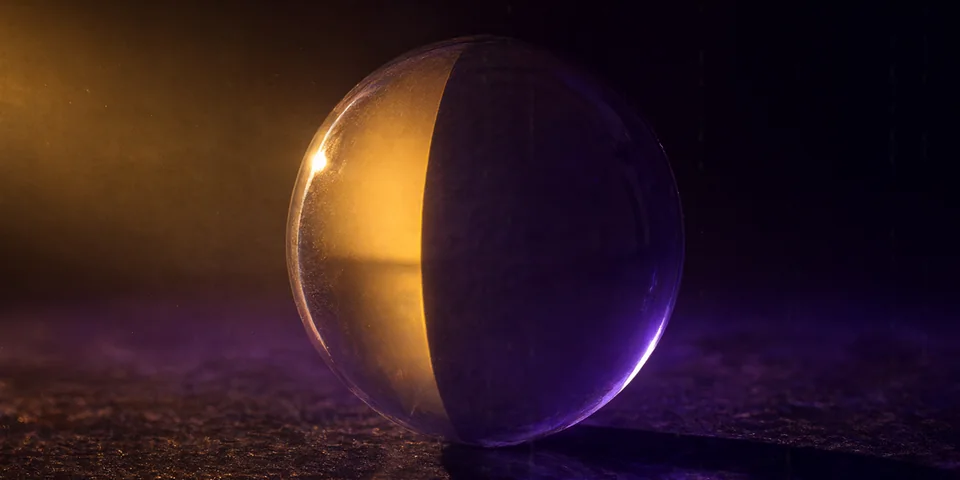 A glass sphere on a dark basalt surface, split by a narrow beam of golden light into a warm amber hemisphere and a violet-shadowed hemisphere.