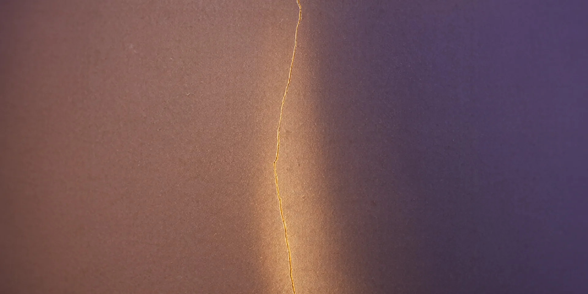 Um vaso de cerâmica mate com uma fina trinca vertical que capta uma linha de luz dourada, o restante em sombra violeta suave.